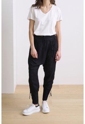 Person trägt ein weißes T-Shirt mit kurzen Ärmeln und V-Ausschnitt, schwarze Jogginghose mit Kordelzug und weiße Sneakers und steht auf einem hellen Holzboden.