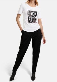 Camicia bianca a maniche corte con un design grafico quadrato in nero e rosso, abbinata a pantaloni neri su misura e stivaletti neri a punta.