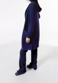 Lång lila cardigan med huva, sidofickor och en avslappnad passform, i kombination med mörka utsvängda jeans och svarta skor. Slät textur.
