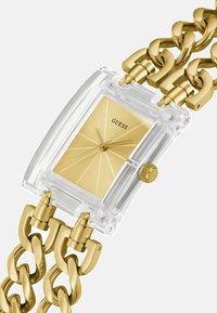 Gennemsigtig rektangulær ur med en guld skive, guld kæderekkearmbånd og et tekstureret, vinklet mønster på urskiven.