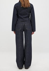 Dunkelblaue Jeans-Outfit mit feinen vertikalen Streifen, bestehend aus einer taillierten Jacke und weit geschnittenen Hosen. Die Nähte sind in einem kontrastierenden Goldton.