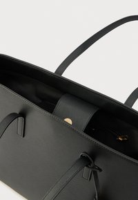 Juodas rankinė-ratukas iš tekstūruotos sintetinės medžiagos, turinti dvi ilgus rankenas, vidinį kišenėlį su užtrauktuku ir apvalią aukso detalę.