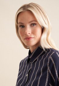 Blusa navy con scollatura a collare, caratterizzata da un motivo a catena in tonalità chiare. Tessuto liscio e morbido con dettagli a bottoni.