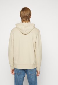 Lys beige hettegenser med snøring, ribbestrikkede mansjetter og en teksturert overflate, kombinert med blå jeans. Enkel design uten synlige logoer.
