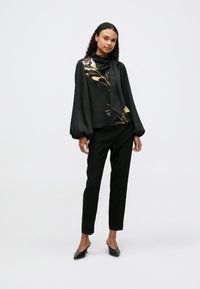Blusa preta com mangas longas e bufantes e um laço no pescoço; apresenta um design floral em rosa e dourado. Combinada com calças pretas justas e saltos altos.