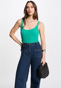 Green ribbed tank top with decorative chain design on straps, paired with high-waisted dark blue wide-leg jeans and a small black handbag.