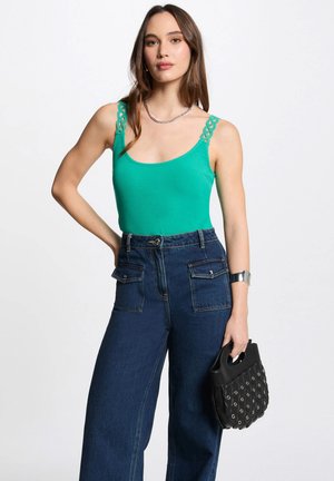 Green ribbed tank top with decorative chain design on straps, paired with high-waisted dark blue wide-leg jeans and a small black handbag.