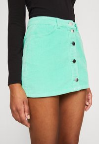 Falda mini de pana verde menta con cintura alta, que cuenta con cinco botones plateados en la parte delantera y bolsillos laterales. Tejido texturizado con ribetes verticales.