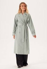 Ljusgrön trenchcoat med bältad midja, dubbelknäppt design och knäppta ärmar, med en elegant och slät yta.