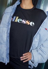 Czarna koszulka z kolorowym, haftowanym logo "ellesse", noszona pod jasnoniebieską pikowaną kurtką o miękkiej teksturze i elastycznych mankietach.