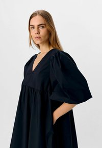 Robe noire avec un décolleté en V profond, des manches ballons, une taille froncée et deux poches latérales. Fabriquée dans un tissu lisse avec une finition mate.