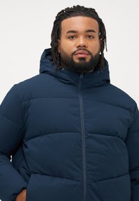 Navy blue puffer jacket with a hood, featuring a quilted design and zipper closure. Smooth texture with no visible patterns or accents.