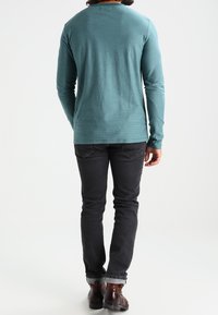 Langärmliges, teal-farbenes Shirt mit Rundhalsausschnitt, kombiniert mit schwarzen Jeans und braunen Stiefeln. Das Shirt hat einen strukturierten Stoff mit feinen Streifen.