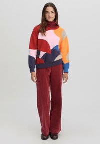 Pull en laine coloré avec des motifs abstraits en rouge, rose, bleu, orange et beige. Associé à un pantalon en velours côtelé large rouge.