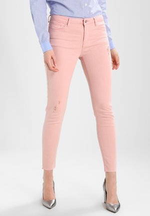 Frau trägt hellrosa schlanke Jeans mit leichter Used-Optik, ein blau gestreiftes Hemd und silberne High Heels, steht vor einem weißen Hintergrund.