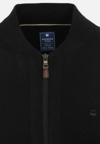 Zwarte zip-up jack met een ribgebreide kraag, metalen rits met leren trekkoord en een merketiket binnenin de kraag. Gladde textuur.