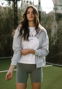 Šedá mikina na zip přes bílém tričku s černým textem, v kombinaci s olivově zelenými sportovními šortkami. V pozadí se nachází zeleň.