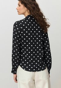 Fekete pöttyös blúz hosszú ujjakkal, lekerekített szegéllyel és gombos mandzsettákkal; az anyag sima, kontrasztos világoskék pöttyökkel.