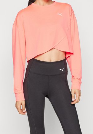 Kobieta ubrana w koralowy długi crop top z długim rękawem i czarne legginsy z wysokim stanem, oba z logo Puma, stojąca na jednolitym tle.