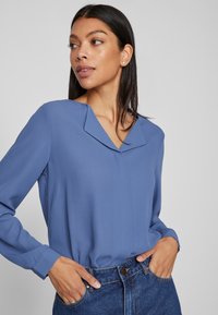Donna che indossa una blusa blu a maniche lunghe con colletto a V e jeans blu, in piedi con le mani nelle tasche, che guarda di lato su uno sfondo semplice.