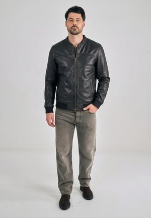 Muž stojící, oblečený v černé kožené bundě, olivově zelené košili, vybledlých šedých džínách a černých botách, na jednoduchém světlém pozadí.