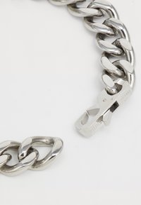 Bracelet en métal argenté avec maillons entrelacés et fermoir portant une gravure de marque. Texture lisse avec une finition brillante.