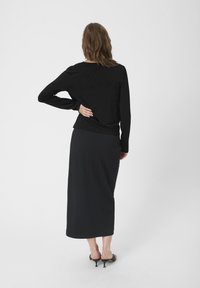Crna majica dugih rukava s opuštenim krojem, u kombinaciji s crnom suknjom do koljena. Model nosi crne sandale i stoji protiv jednolične pozadine.