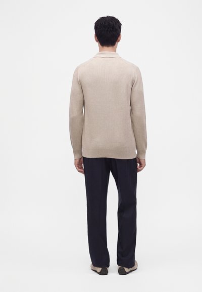 Homme debout de dos, portant un pull beige à manches longues, un pantalon foncé et des chaussures beiges sur un fond blanc uni.