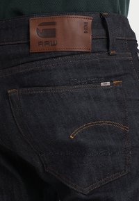 Tmavě modré džíny z denimu s koženou nášivkou loga, oranžovým prošíváním a jednou zadní kapsou s malou značkovou cedulkou. Viditelný je texturovaný detail látky.