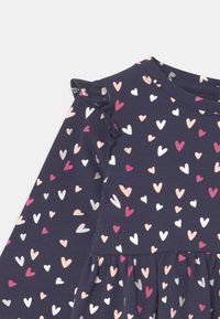 Tmavě modré šaty s dlouhým rukávem a volánkovým detailem na rameni, zdobené rozptýlenými srdíčky v růžové, bílé a stříbrné barvě. Měkká bavlněná látka, vícedílný střih.