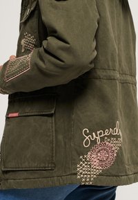 Olijfgroene jas met geborduurde accenten, waaronder bloemmotieven en het woord "Superdry." Gestructureerde stof met meerdere zakken.