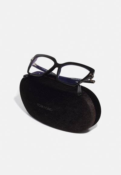 Gafas de Tom Ford negras con montura gruesa y lentes transparentes, colocadas sobre un estuche redondeado de terciopelo negro con el nombre de la marca en relieve.