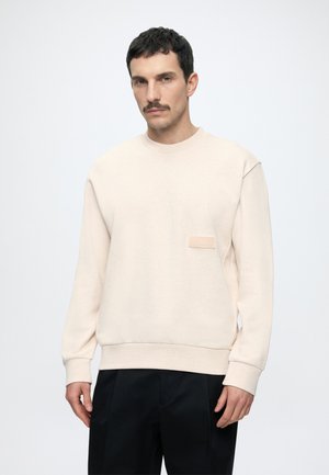 Homme aux cheveux courts et foncés avec moustache, portant un sweat-shirt beige clair avec un petit écusson « BOSS » sur la poitrine et un pantalon noir.