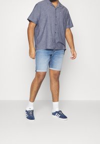 Camicia blu navy a maniche corte con bottoni, pantaloni shorts in denim azzurro arrotolati alle estremità, e sneakers in suede blu con strisce bianche. Calzini bianchi alla caviglia inclusi.