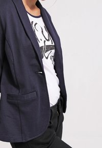 Merimiessininen bleiseri, jossa on yksirivinen nappi, lovikaulukset ja päällystetyt taskut. Pehmeä, kevyt kangas, jossa on sileä pinta. Päällä graafinen t-paita.