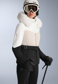 Skijaška jakna s bijelim i bež gornjim dijelom, crnim donjim dijelom, kapuljačom obloženom krznom i kviltanom teksturom. Uključuje rukavice i skijaške štapove.