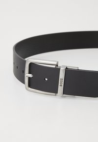 Curea din piele neagră, cu o cataramă metalică argintie gravată cu logo, ajustabilă cu găuri, textură netedă și un design clasic.