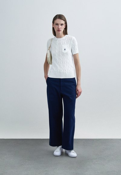 Λευκό κοντομάνικο πουλόβερ με πλεκτό σχέδιο και τσέπη, συνδυασμένο με μπλε σκούρο φαρδύ παντελόνι και λευκά παπούτσια, δημιουργώντας ένα καθημερινό σύνολο.