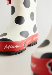 Weiße Gummiregenstiefel mit großen schwarzen Punkten, roten Sohlen und einer Schriftzug-Detaillierung, die "Minnie" zusammen mit einem Minnie-Maus-Icon liest.
