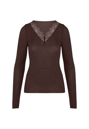 Vrouw met blond haar die een aangesloten lange-mouwen bruin shirt en bijpassende bruine broek draagt, met haar handen gekruist achter haar rug.