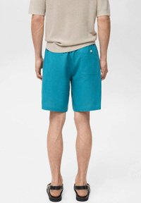 Pantaloni corti turchesi in cotone con una vestibilità comoda, caratterizzati da una vita elastica e una tasca posteriore con dettaglio a bottoni. Indossati con una camicia beige.