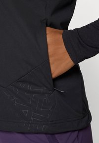 Black athletic top with a textured black panel and a side pocket. Long sleeves with a dark purple accent at the bottom.