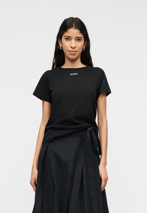 Hosszú sötét hajú nő fekete, rövid ujjú felsőt visel „PINKO” felirattal és fekete, derékban megkötött wrap szoknyát, egy egyszerű háttér előtt állva.