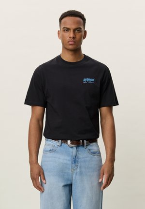 Homme portant un t-shirt noir « Prince Les Deux » et un jean bleu clair, debout devant un fond beige uni.