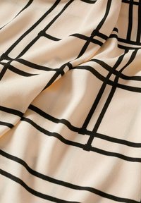 Tissu beige à motif quadrillé noir et texture lisse, présentant des lignes horizontales et verticales de largeurs variables.