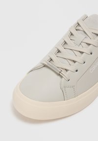 Light gray sneaker with a smooth leather upper, round toe, and beige rubber sole. Features flat laces and a metallic logo accent.