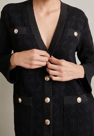 Hände, die eine schwarze, strukturierte Strickjacke mit goldfarbenen Knöpfen und gerippten Abschlüssen an Taschen und Ausschnitt zuknöpfen.