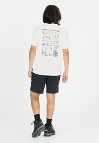 Vaalea beige t-paita, jossa on mustia graafisia kuvastoja selässä, yhdistetty mustiin shortseihin ja karkeisiin mustiin jalkineisiin. Valkoinen tausta.