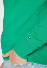 Felpa verde realizzata in morbido tessuto, con polsini a coste e un piccolo etichetta con logo rosso, bianco e blu sulla manica.