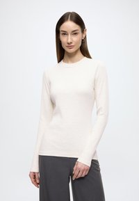 Langärmliges cremefarbenes, geripptes Shirt mit rundem Ausschnitt, figurbetonter Silhouette und weicher Textur, kombiniert mit dunkelgrauen Hosen.